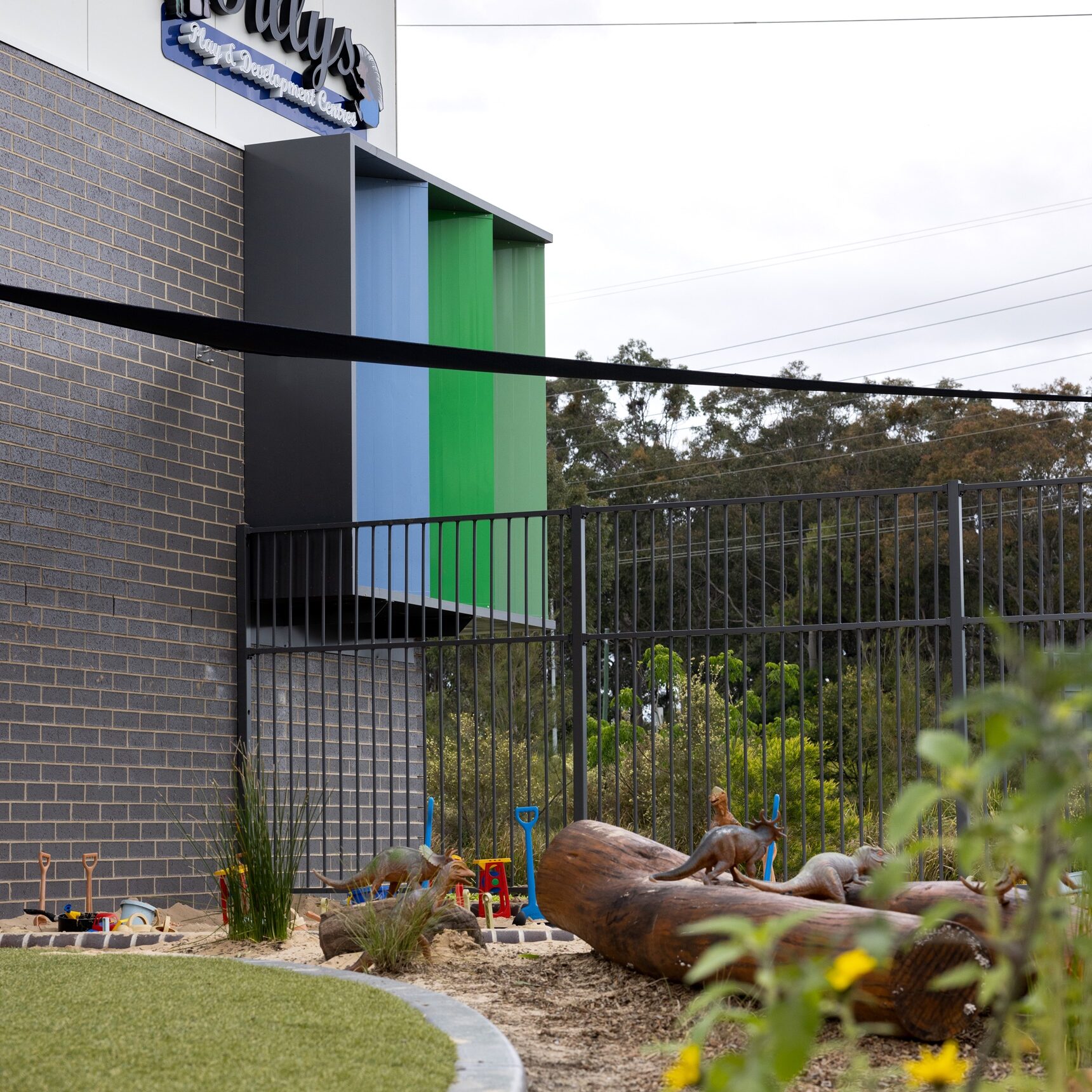 Modern building exterior with colorful panels in blue, green, and black. A "Tillys" logo sign is on the wall. Foreground features landscaped garden with rocks and plants. Overcast sky.
