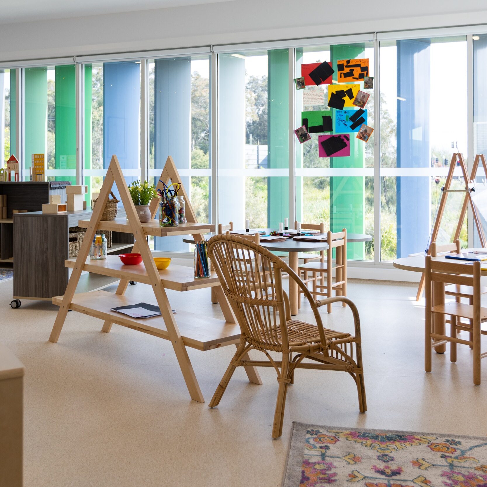 Spacious art room with colorful vertical window blinds. Wooden easels, wicker chairs, and a floral-patterned rug create a bright, creative atmosphere.
