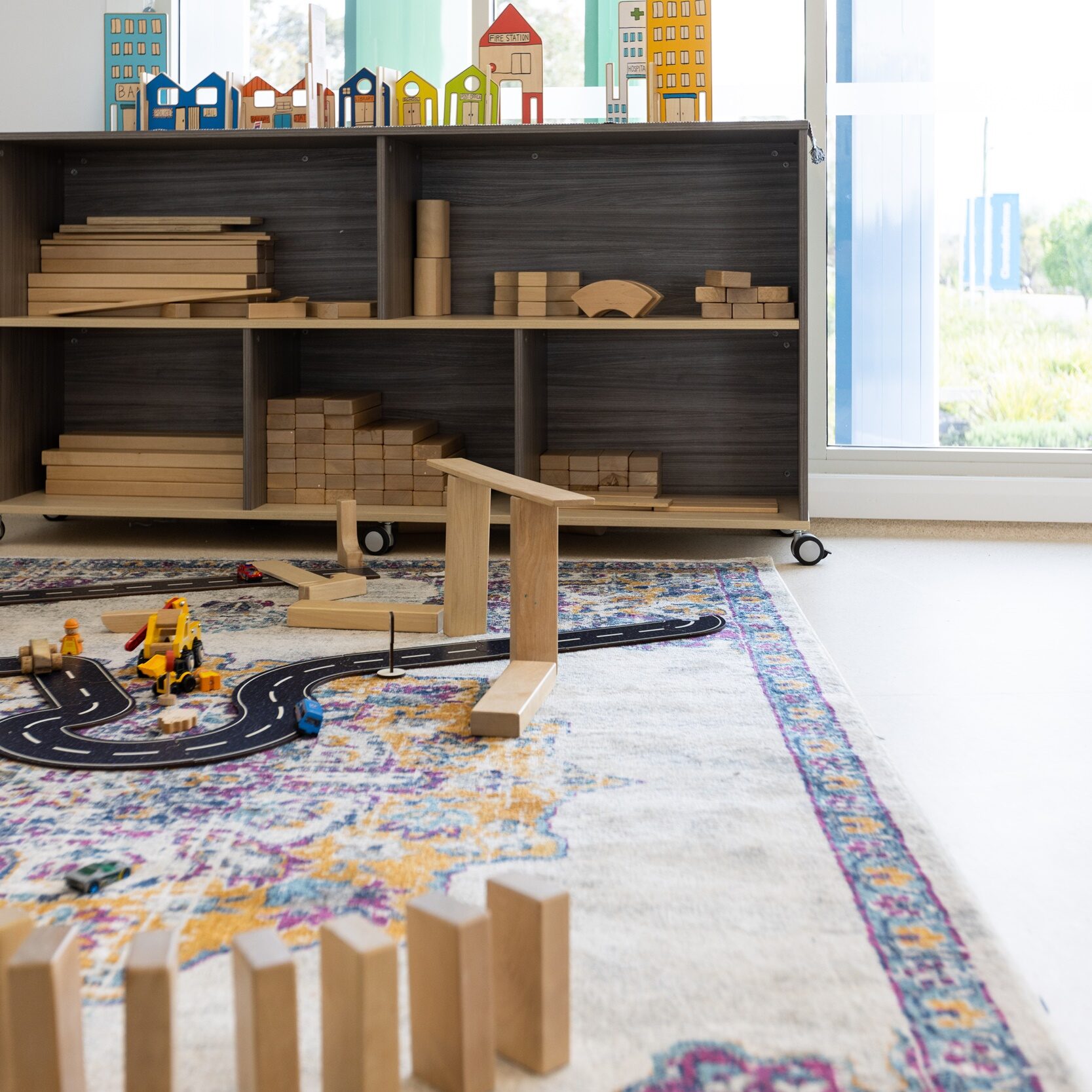 A cozy playroom with wooden blocks and toys scattered purposefully on a colorful rug. A shelf holds toy houses by a large window, creating a bright, playful atmosphere.