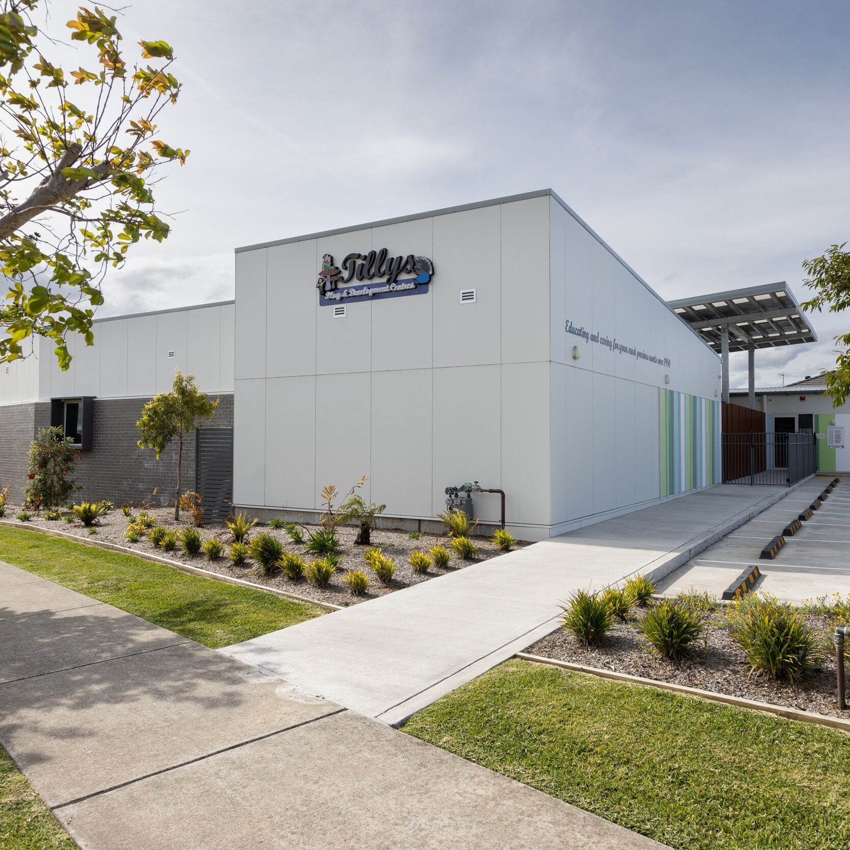 Modern white building with" Tillys Child care" logo sign, surrounded by neatly landscaped greenery and a paved walkway under a partly cloudy sky.
