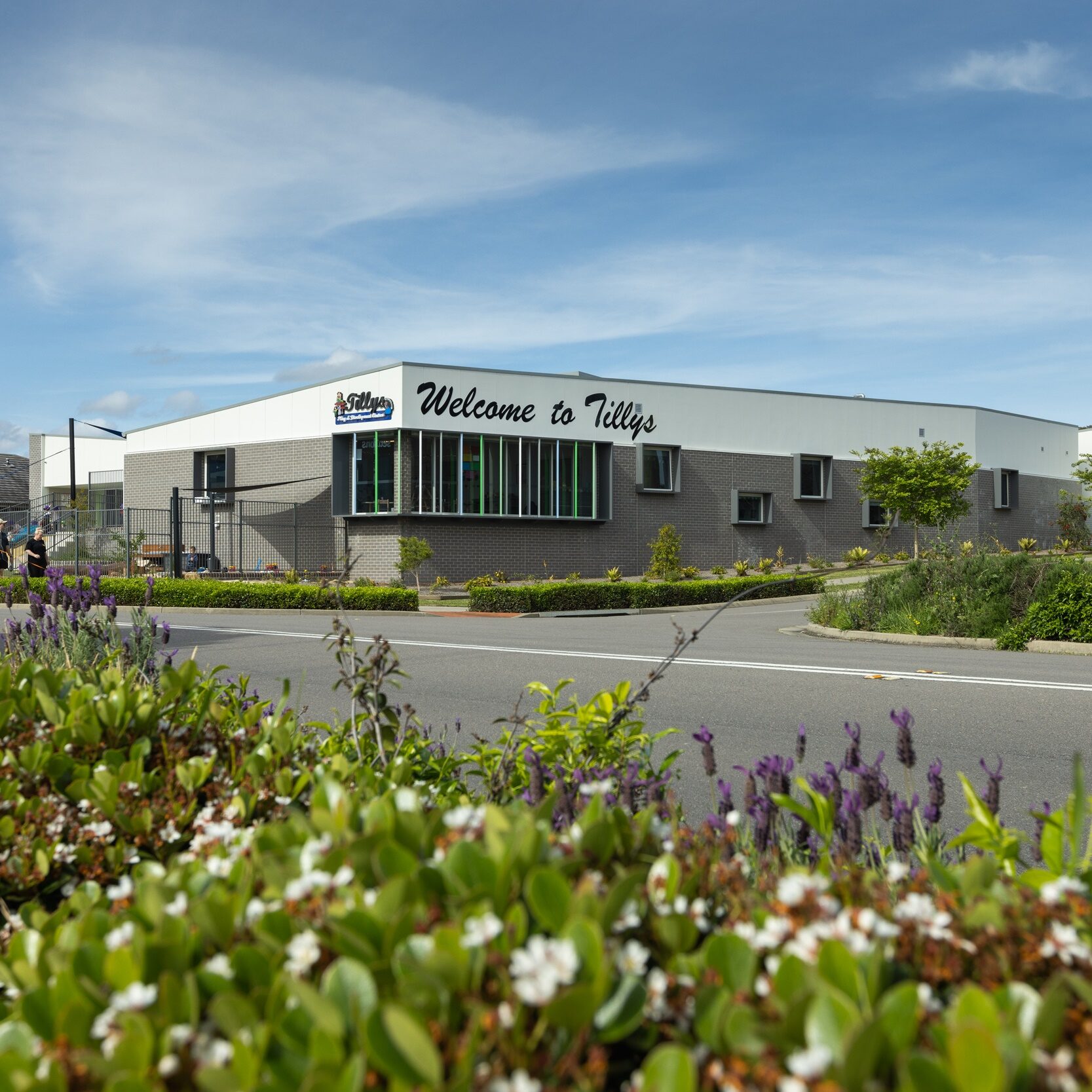 Modern building with "Welcome to Tillys" sign, surrounded by lush greenery and flowers on a sunny day with blue skies and scattered clouds. Peaceful setting.