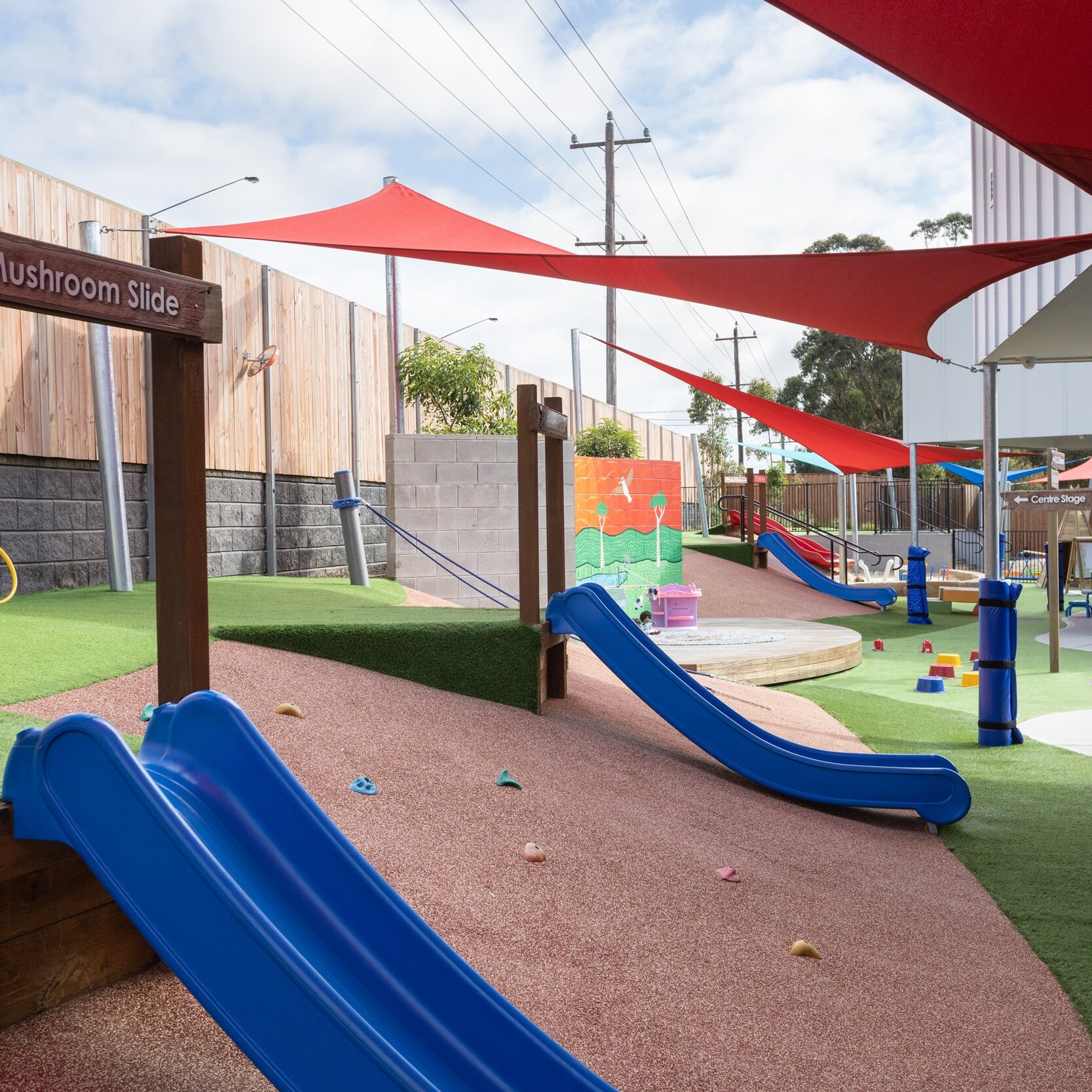 Outdoor playground with blue slides, grassy areas, and red sunshades. Wooden fence and playful climbing rocks create a vibrant, joyful atmosphere.