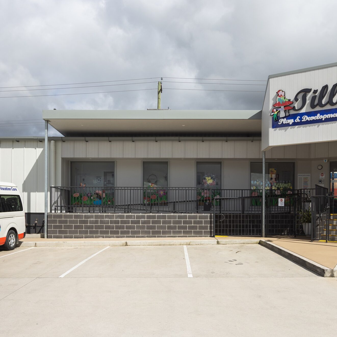 Front view of Tillys Play & Development Centre, with a white mini bus parked outside. The building is gray with a large sign, under a cloudy sky.
