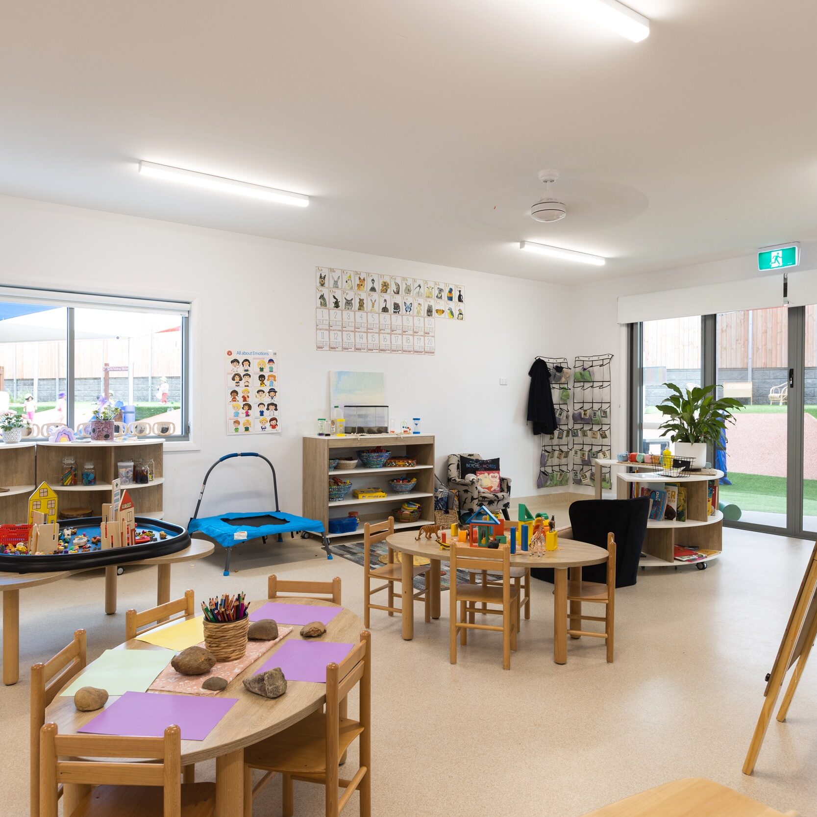 Bright, spacious classroom with round tables, small chairs, art supplies, and shelves. Large windows offer natural light, creating a welcoming, educational atmosphere.