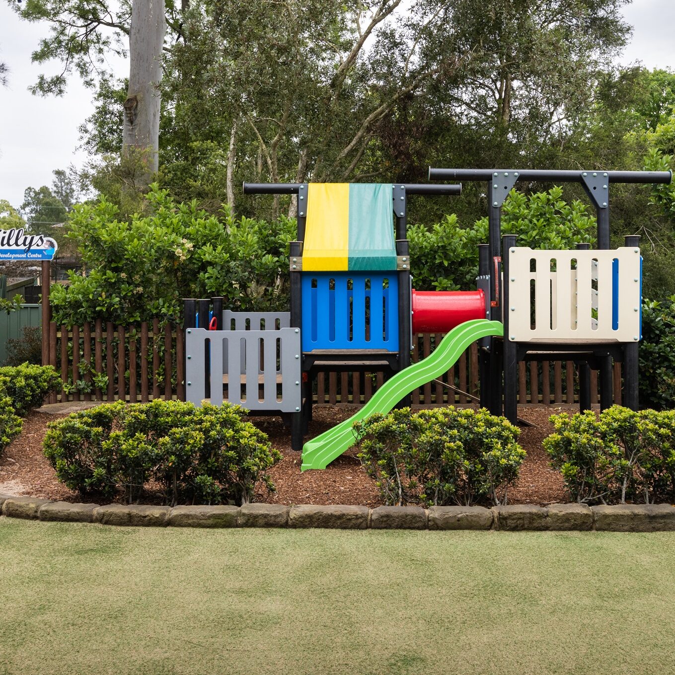 Playground with colorful slides and climbing structures surrounded by lush natural greenery. A wooden logo sign with 'Tillys' enhances the playful, inviting atmosphere.