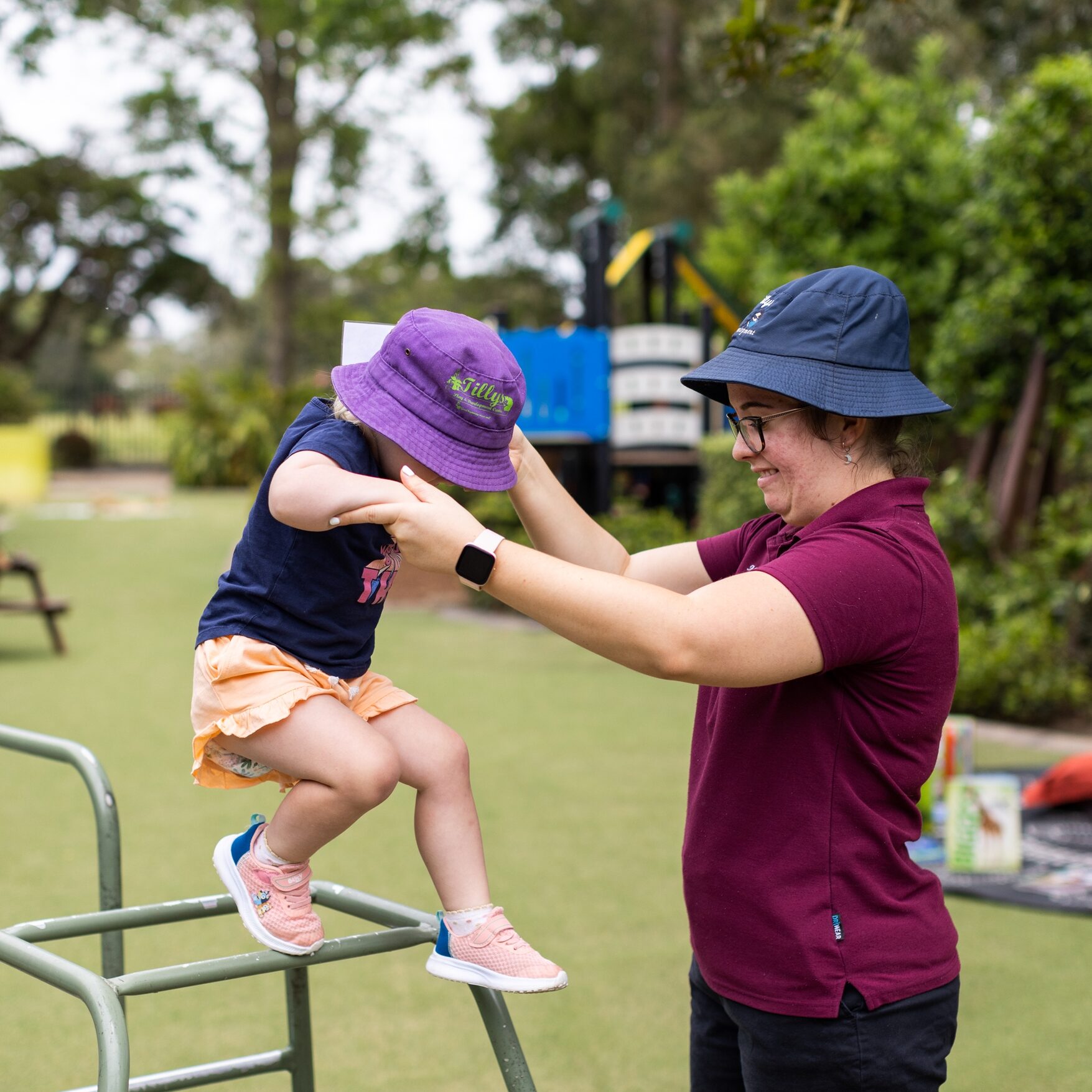 A child in a purple hat sits atop playground climbing bars, reaching for support from an adult teacher wearing a navy hat. They are outdoors, surrounded by trees.