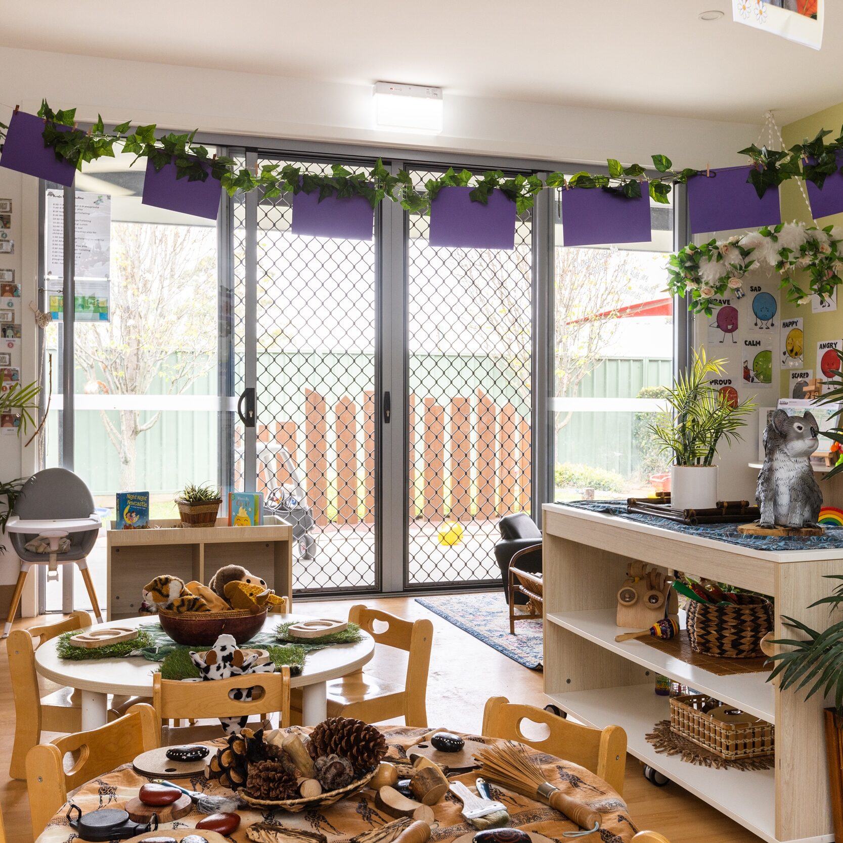 Bright classroom with wooden tables and chairs arranged for play. Plants, colorful paper, and children's artwork create a lively, welcoming atmosphere.