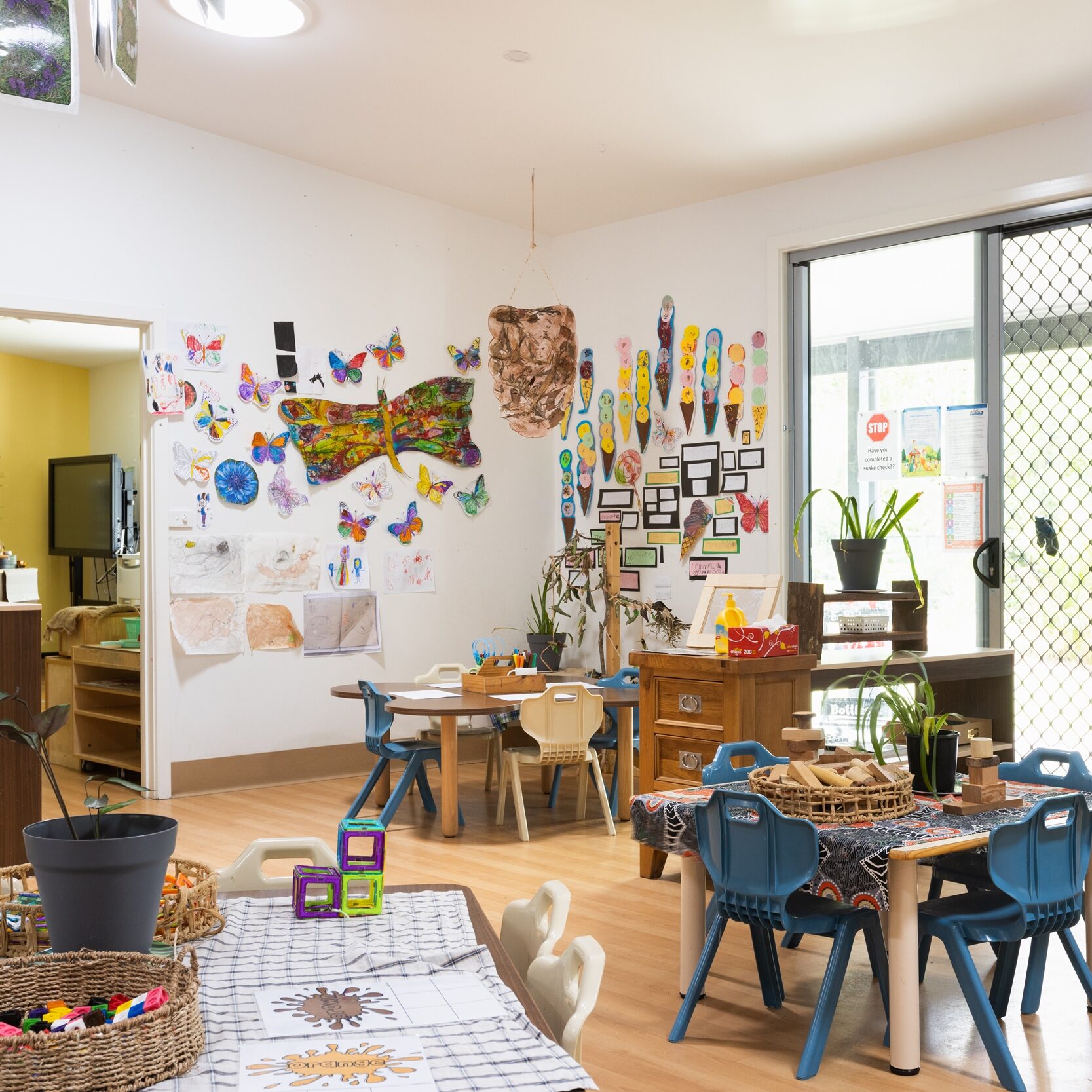 Bright, inviting classroom with colorful children's artwork on walls, small tables with blue chairs, and light streaming through large windows. Creative and lively ambiance.