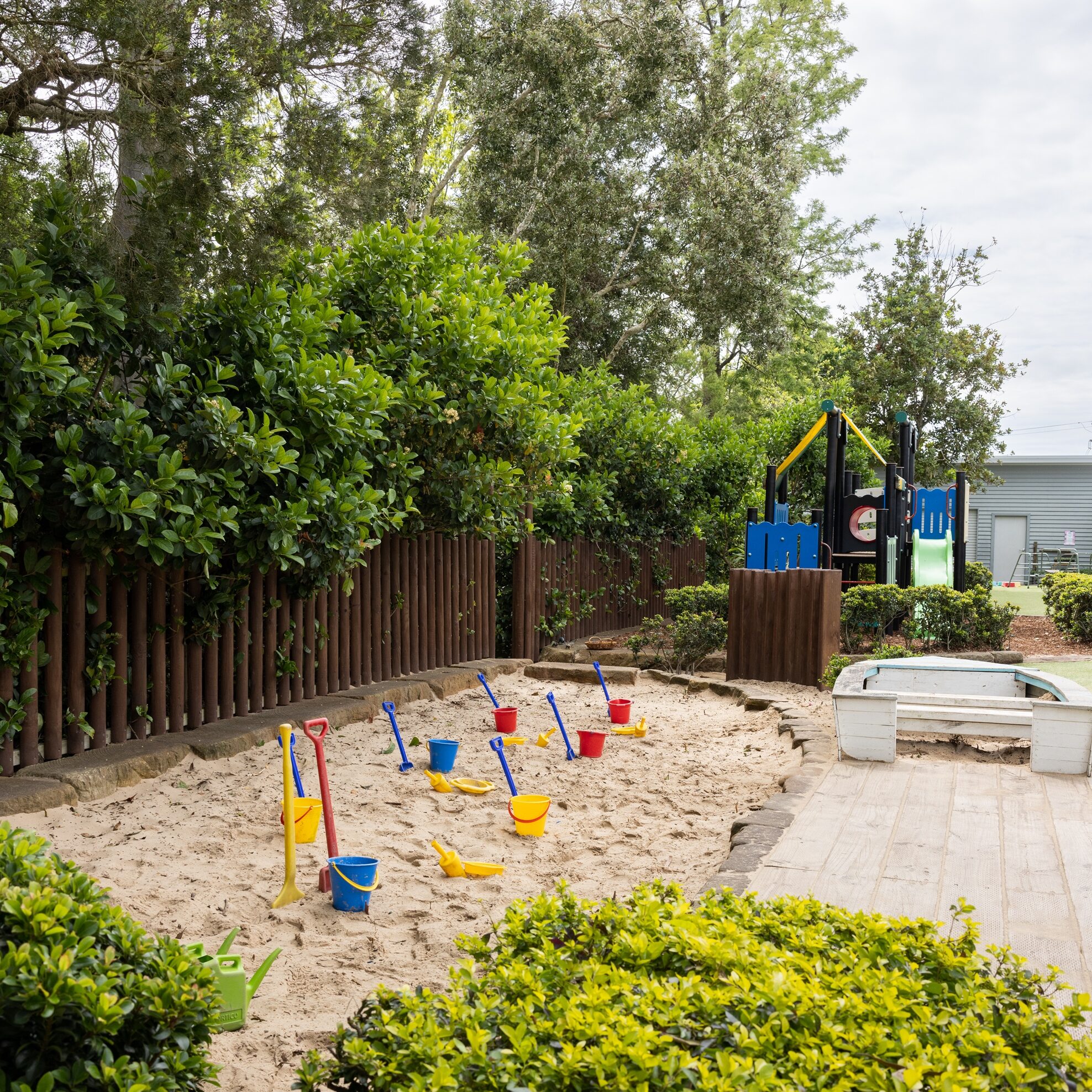Children's large sandpit surrounded by lush natural greenery, filled with colorful buckets and spades. Nearby, a wooden play structure under a cloudy sky.