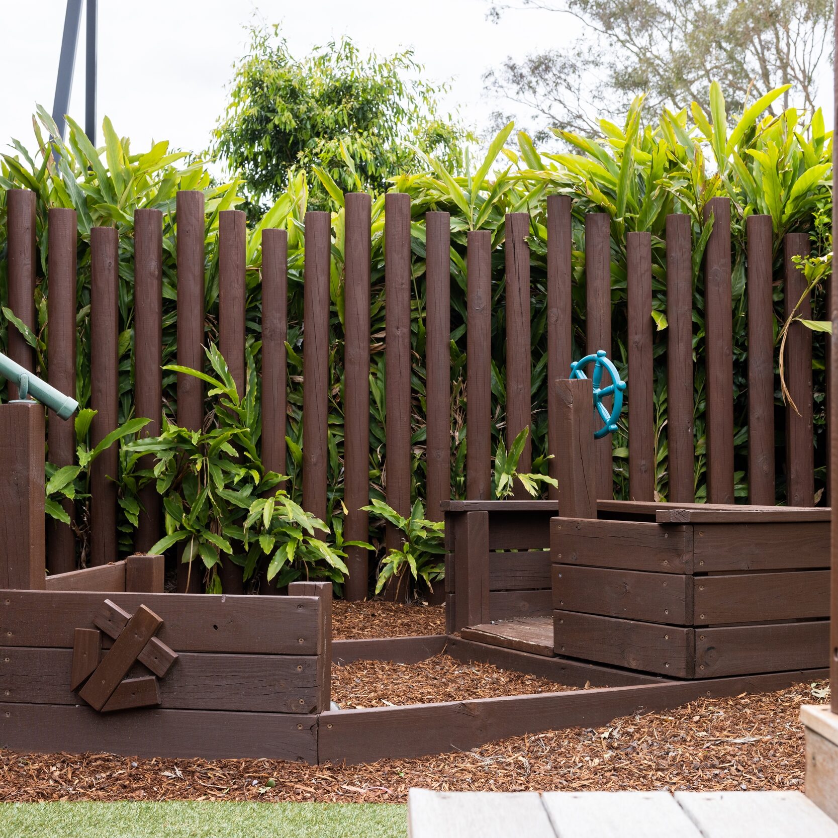 Wooden boat playground with a sandbox surrounded by green plants and a wooden fence. The setting feels playful and natural.