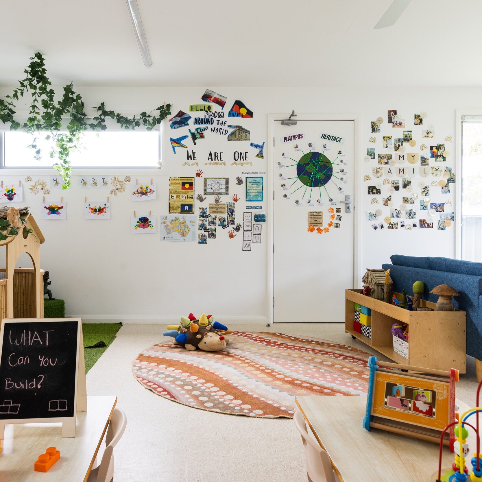 Bright, colorful classroom with a cozy rug, blue sofa, and educational toys. Walls display children's artwork and plants, creating an inviting atmosphere.