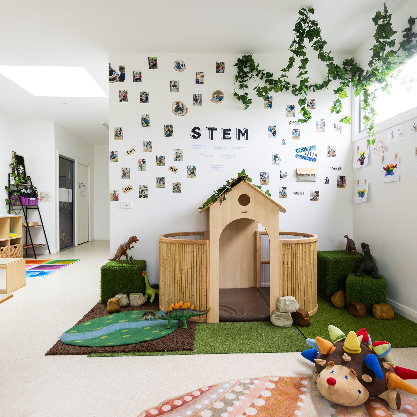 Bright, welcoming playroom with a STEM-themed wall display. It has a cozy wooden playhouse, lush artificial grass, hanging greenery, and colorful toys creating a playful, imaginative atmosphere.