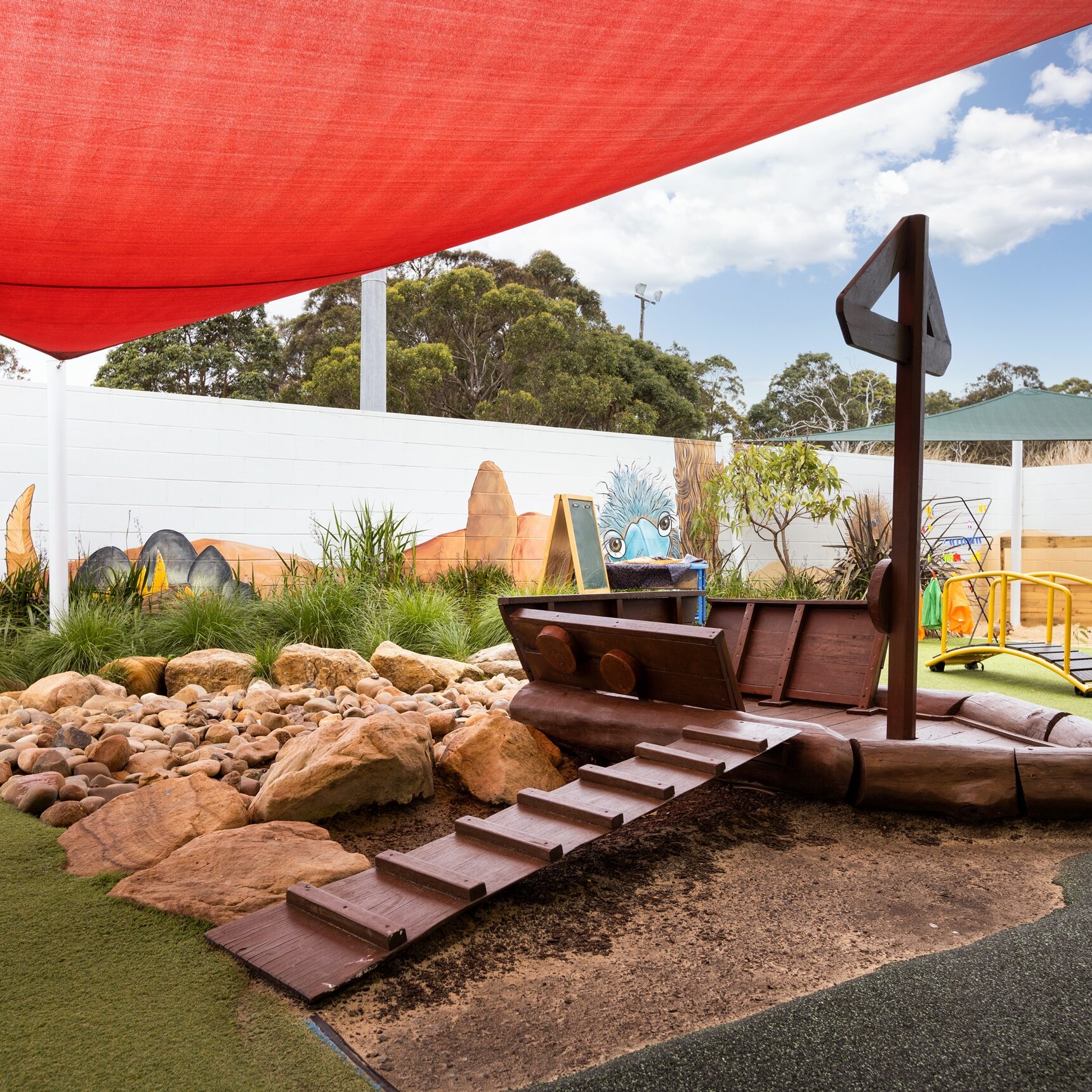 Children's playground with a wooden pirate ship structure on soft ground. A wall mural shows a desert scene with animals. Red sunshade overhead.