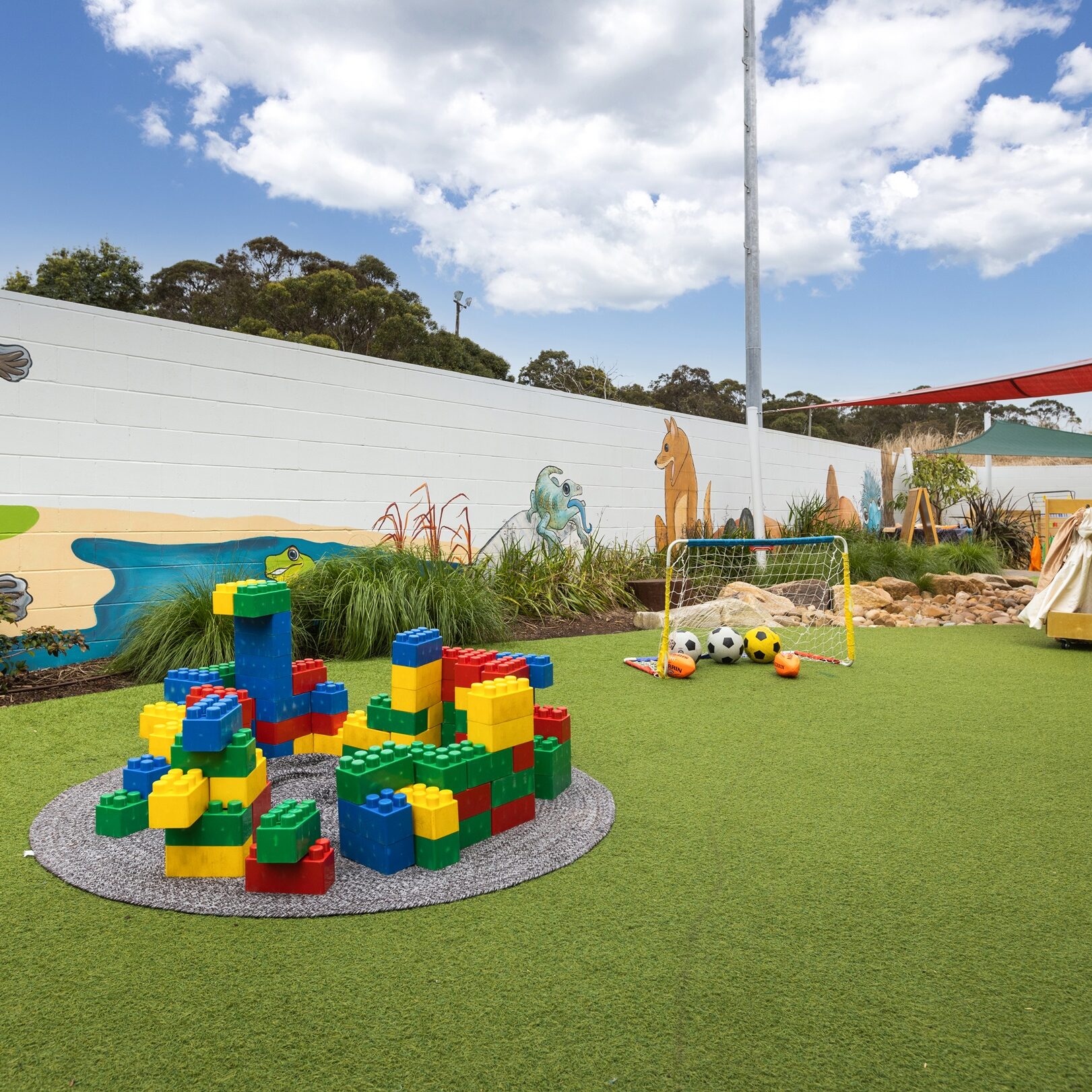 A vibrant playground with colorful foam blocks on artificial grass, surrounded by a wall with animal murals. Bright blue sky and red shade add cheerfulness.