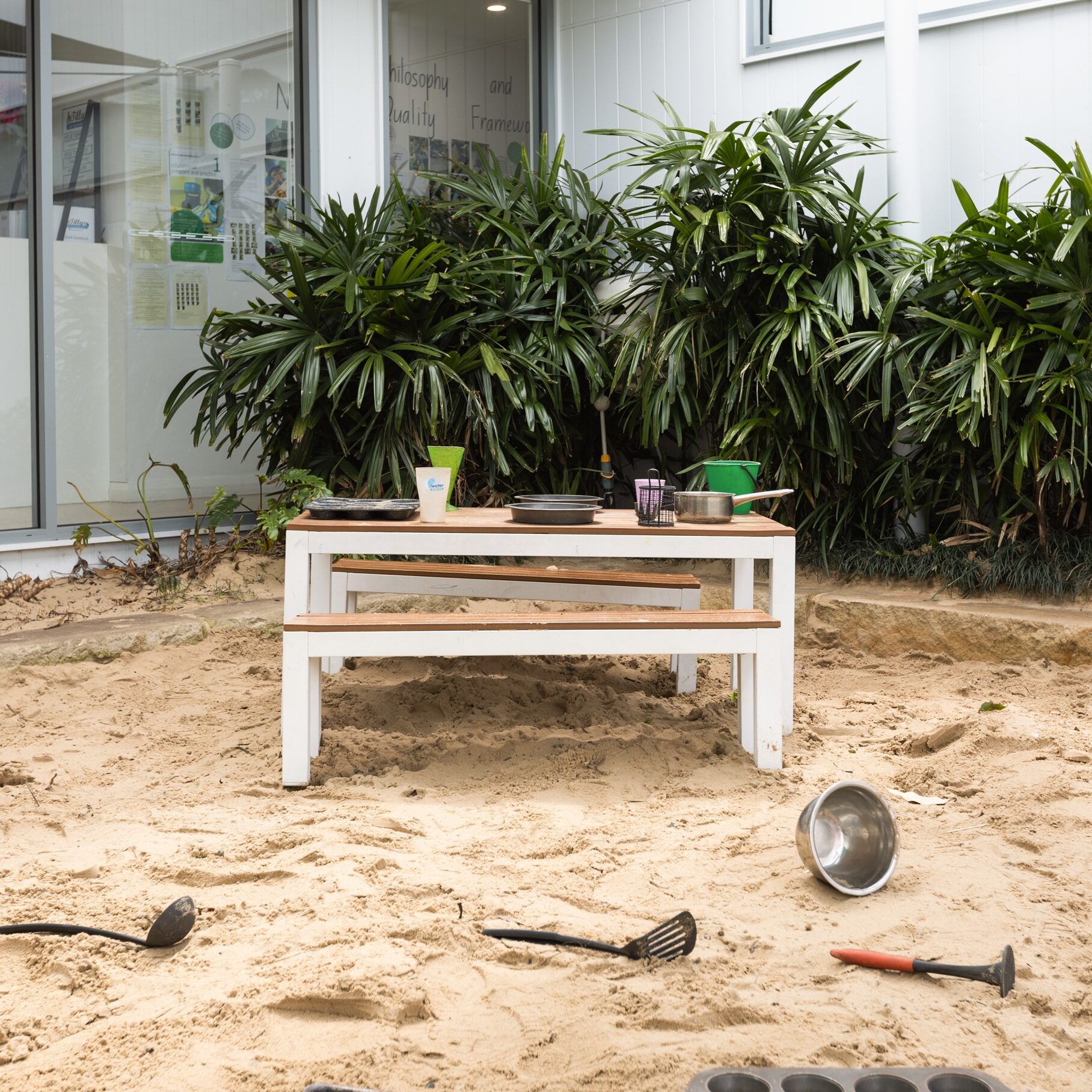 Outdoor sandpit kitchen with white table, surrounded by lush green plants. Baking trays, ladles, and utensils scattered in the sand, evoking playfulness.