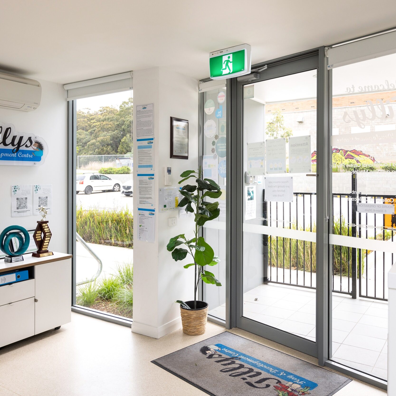 Bright office entrance with glass doors. Trophies line a cabinet under a "Tillys" sign. Potted plant and safety notices create an inviting, professional atmosphere.