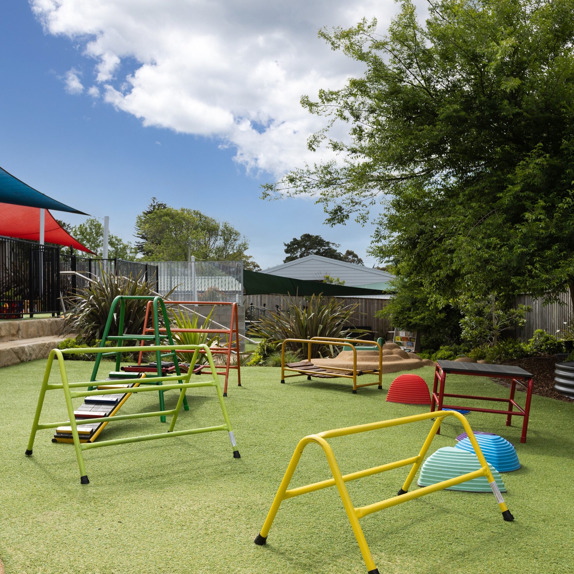 Playground with climbing frames, slides, and tunnels on green grass. Surrounded by trees and bushes under a bright blue sky with fluffy clouds.