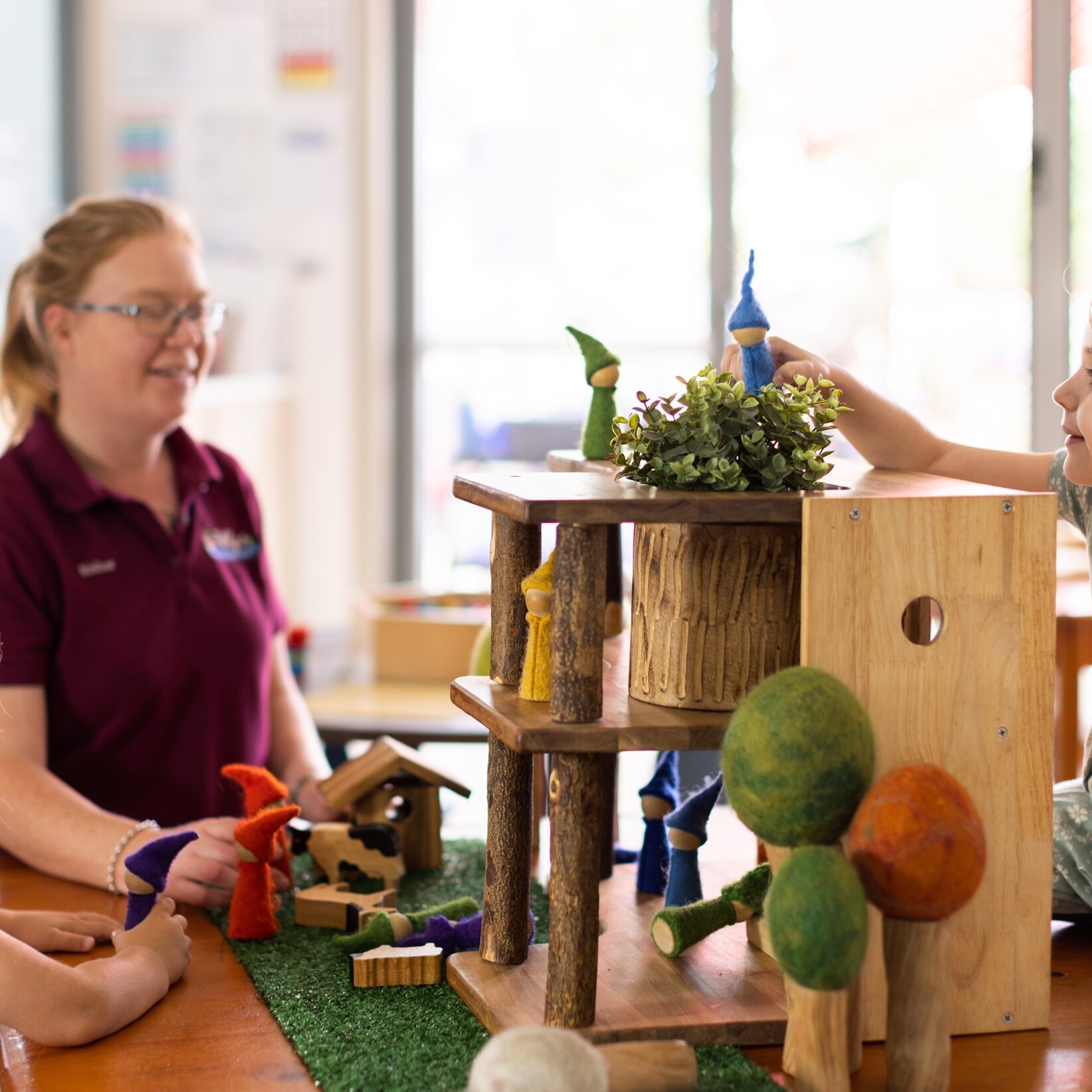 Two young girls and a teacher engage with a wooden playset on a table. The scene is bright, fostering creativity and imagination in a playful setting.