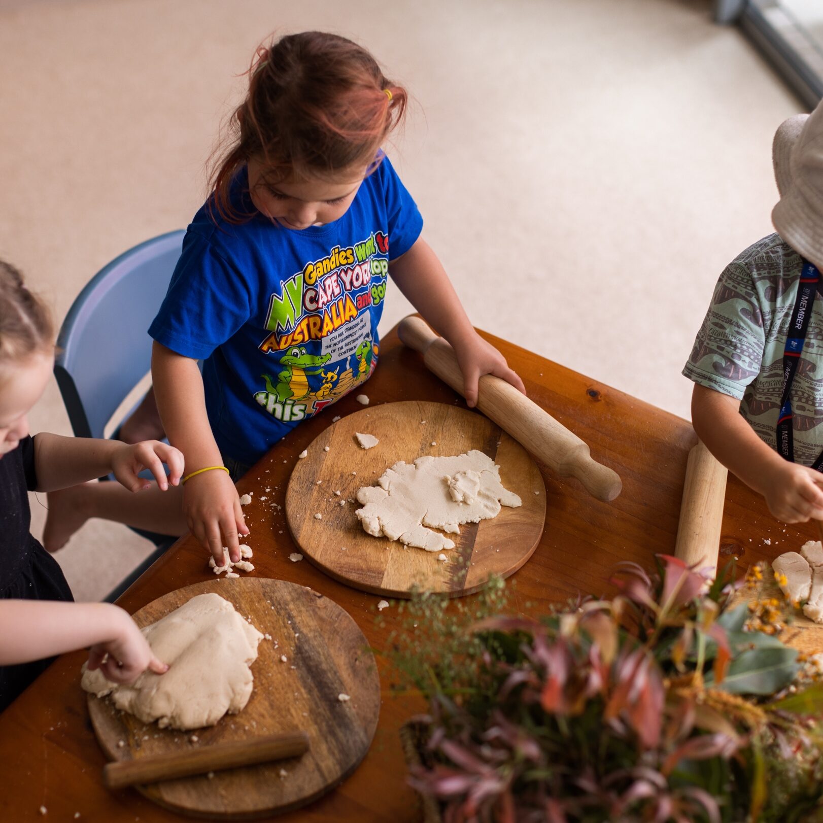 Three children are engaged in play, rolling dough at a wooden table. Each child has a rolling pin, and there's a plant centerpiece, creating a creative and focused atmosphere.