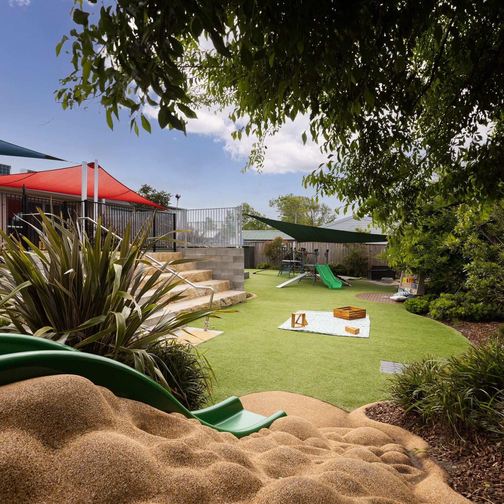 Outdoor playground with green grass, slides, sandpit, and shade sails on a sunny day. Lush plants and trees provide a vibrant, inviting atmosphere.