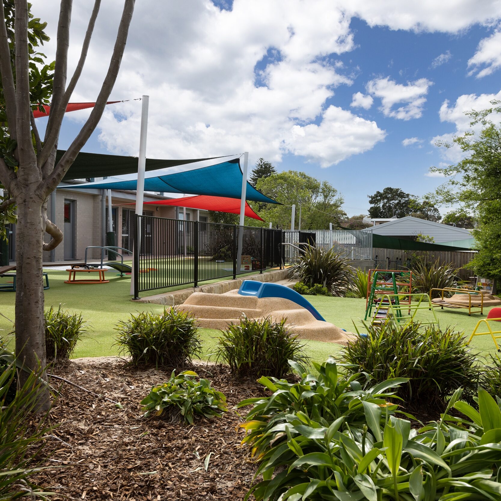 Playground with colorful shade sails, slide, and lush greenery. Bright, sunny day with scattered clouds, conveying a lively and inviting atmosphere.