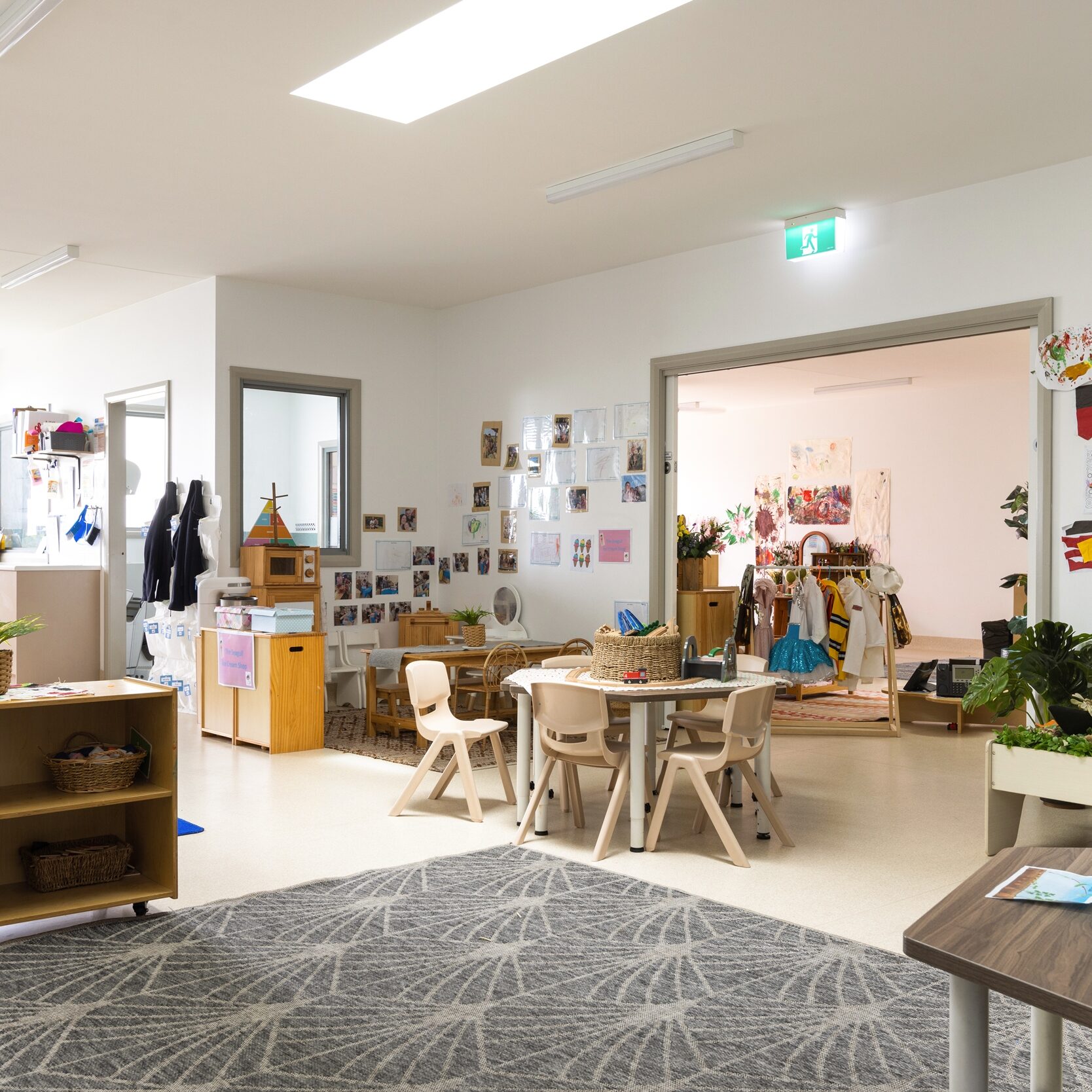 Spacious classroom with scattered tables and chairs. Walls adorned with colorful art. Shelves hold plants and toys, creating a warm, inviting atmosphere.
