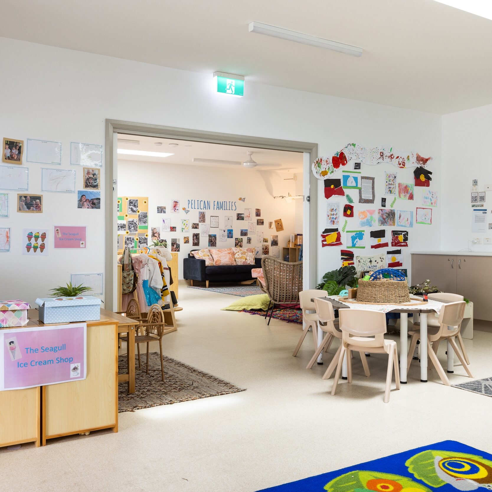 Bright classroom with colorful artwork on walls, small wooden tables and chairs, and a play area with a vibrant rug. Inviting and cheerful ambiance.
