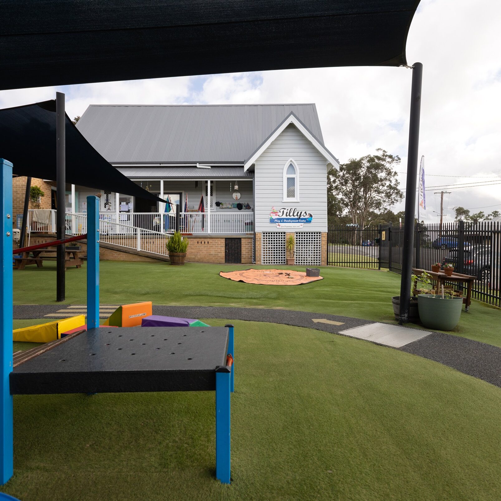 A playground with colorful play equipment on an artificial grass surface. A small building with a gray roof and white facade is in the background with a Tillys logo on the front. It is surrounded by trees and a black fence. The atmosphere is overcast