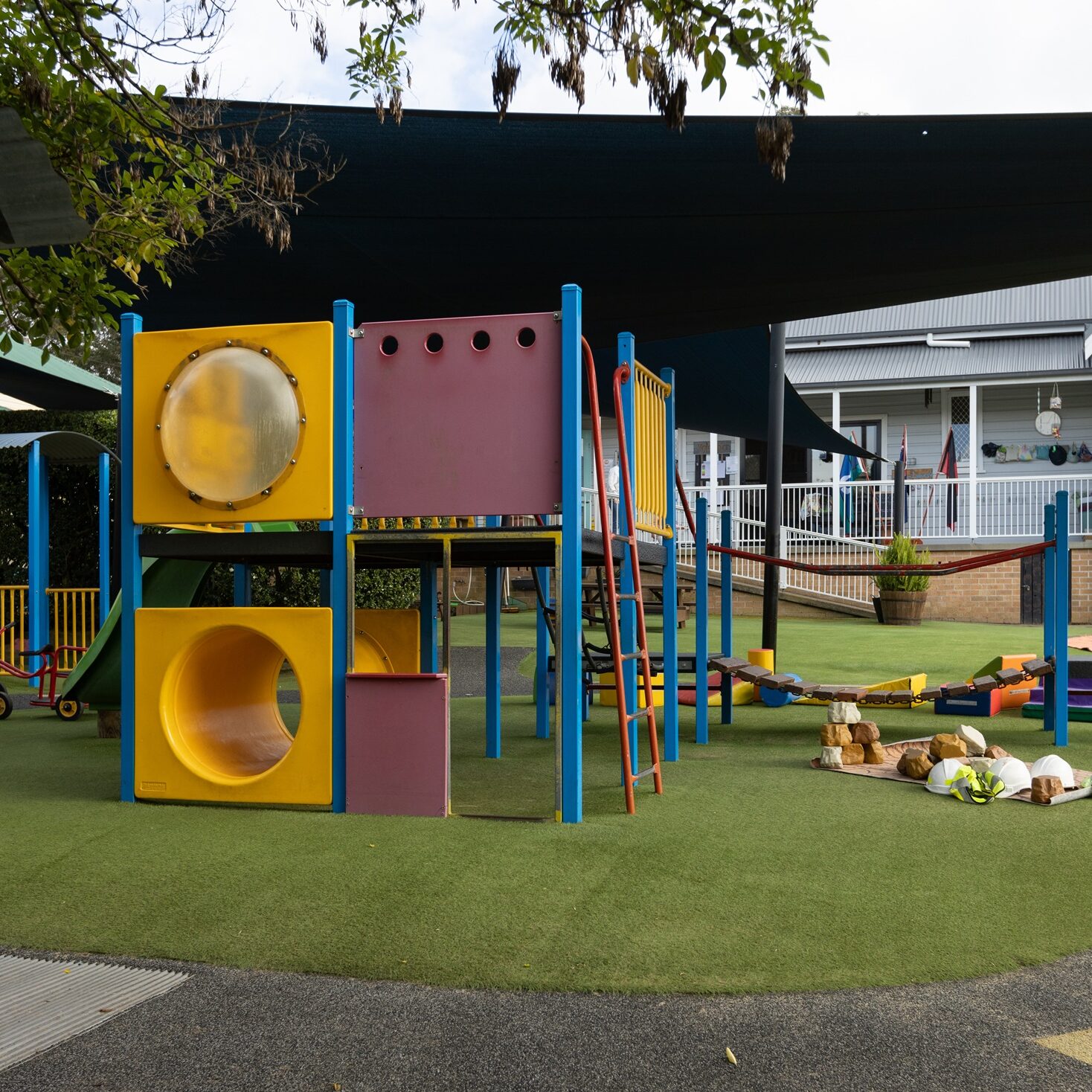 Playground with colorful climbing structures featuring yellow tunnels and blue poles. A bike track with arrows wraps around the climbing structure. Nearby, is a white building with the logo 'Tillys Play and Development Centre'.
