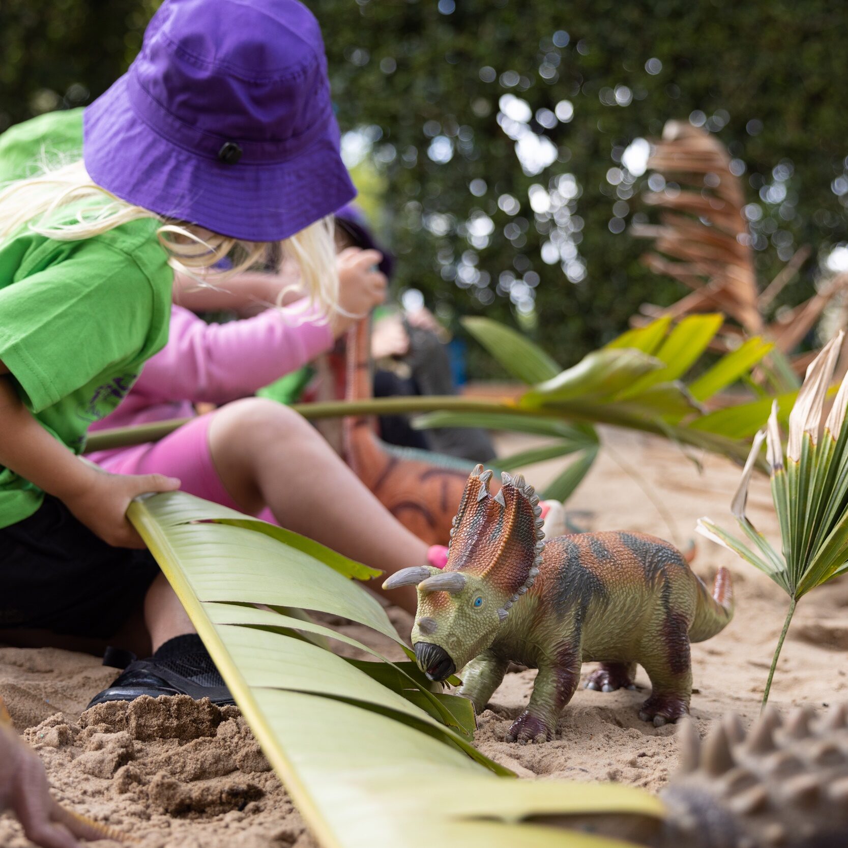 Children sit in a sandpit with natural items such as leaves and large branches, along with toy dinosaur figurines. The scene is sunny, colourful and inviting for children to play in
