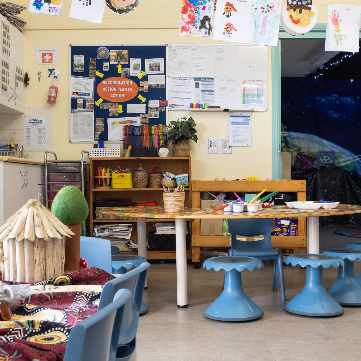 Cozy classroom filled with round tables, blue chairs, wall art, crafts, and educational displays. Bright and inviting atmosphere with creative decor.