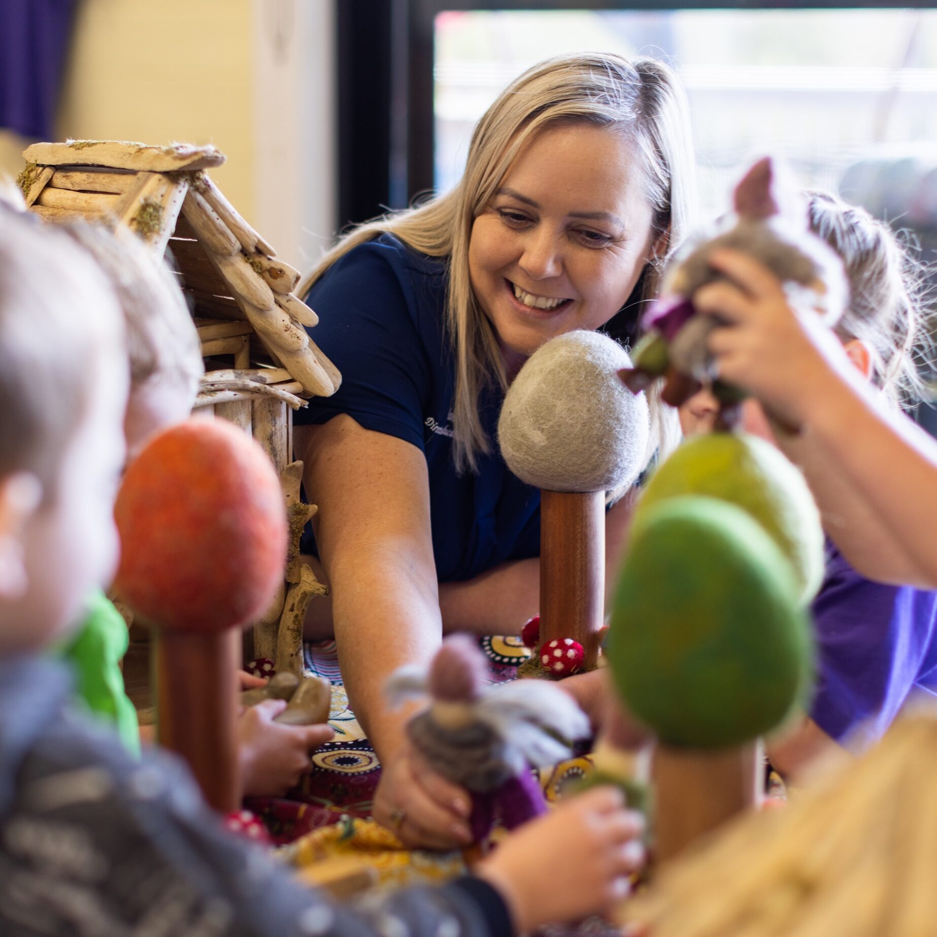 A teacher smiles and interacts with young children playing at a table with colorful toy trees and structures, evoking a playful and engaging atmosphere.