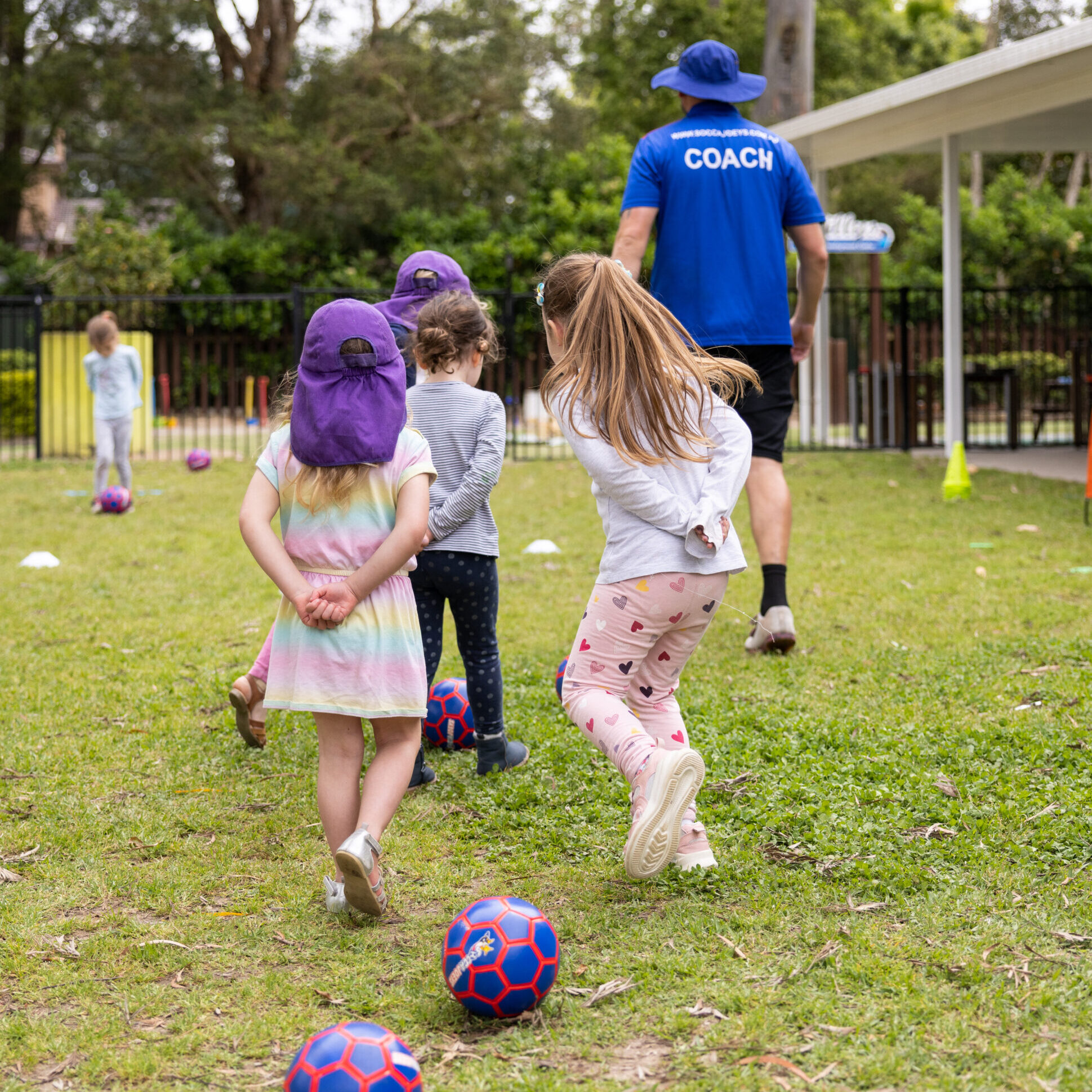 Children follow a coach wearing a blue shirt in a grassy area with trees. Two colorful soccer balls and cones are scattered on the field, suggesting a playful, energetic mood.