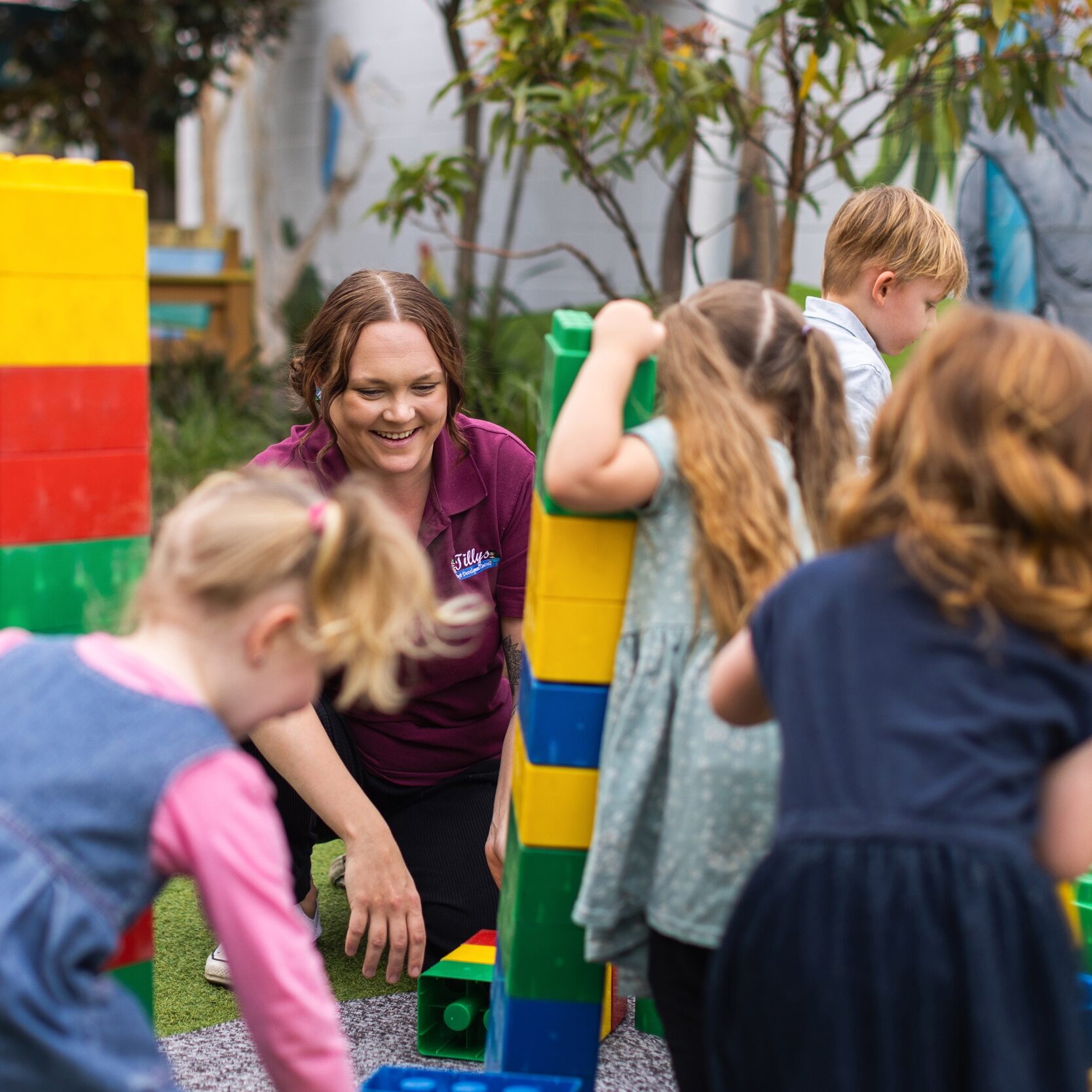 Children play with large, colorful toy blocks outside, supervised by a smiling teacher. The scene is lively and cheerful, with kids actively engaged.