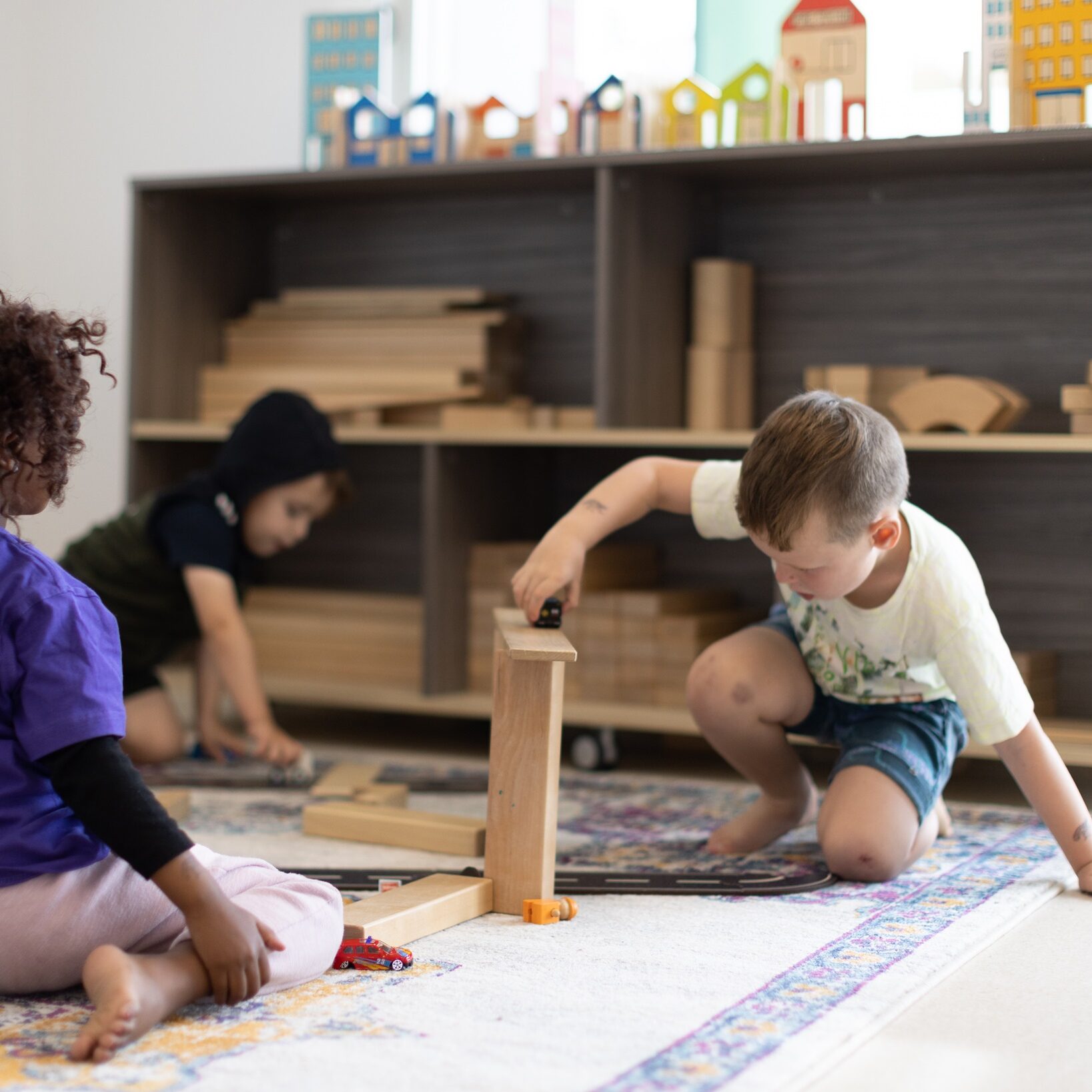Three children play with wooden blocks on a carpeted floor. They are focused and engaged, with shelves of more blocks and colorful houses behind them.
