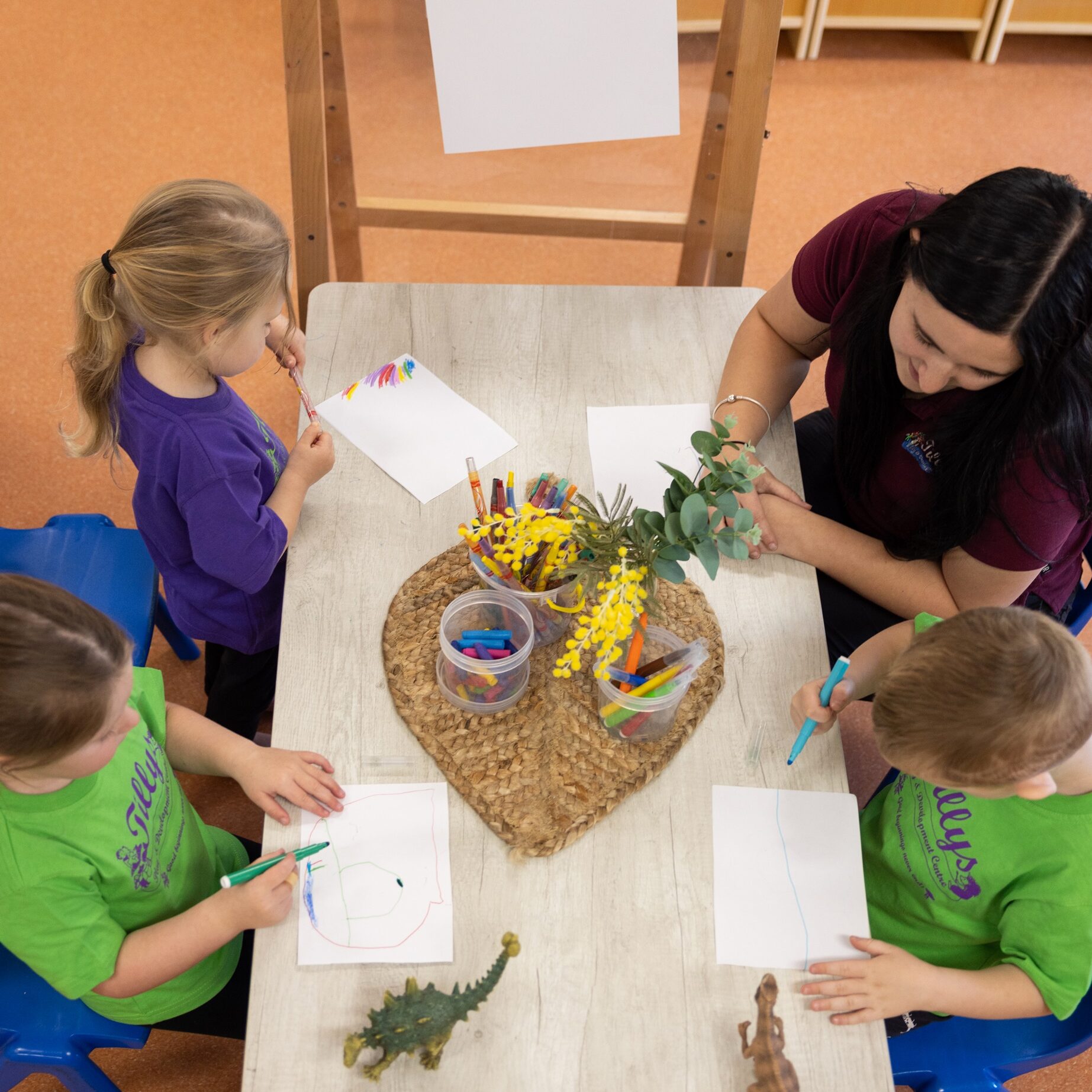 Children and a teacher sit around a table engaged in drawing activities. A heart-shaped basket with plants and toy dinosaurs decorates the table, creating a lively atmosphere.