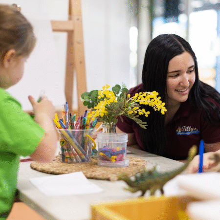 Children engaged in drawing at a table with colorful pencils and paper. A teacher smiles, observing, with a vase of yellow flowers nearby. Bright and cheerful setting.
