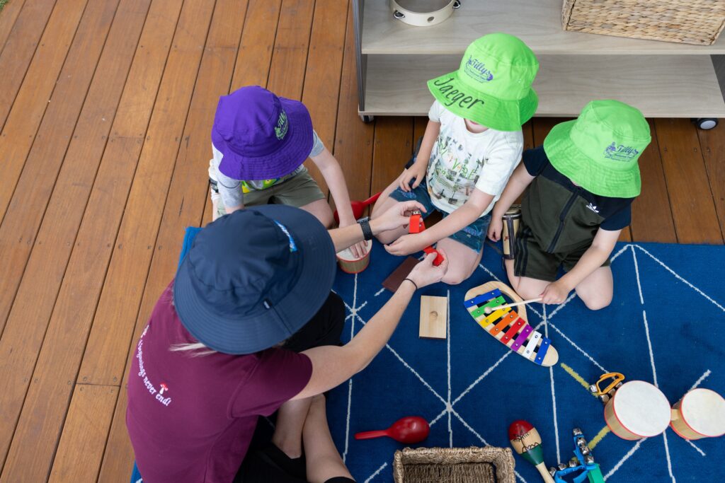 Tillys A Tillys teacher and three children, seated on a blue patterned rug, play with colourful musical instruments. The children wear bright hats, and the atmosphere is playful.