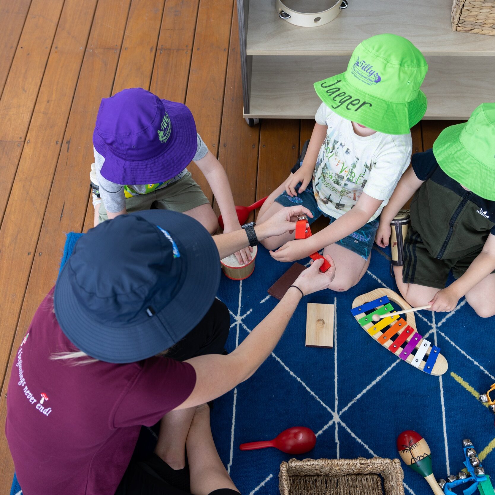 A Tillys teacher and three children, seated on a blue patterned rug, play with colourful musical instruments. The children wear bright hats, and the atmosphere is playful.