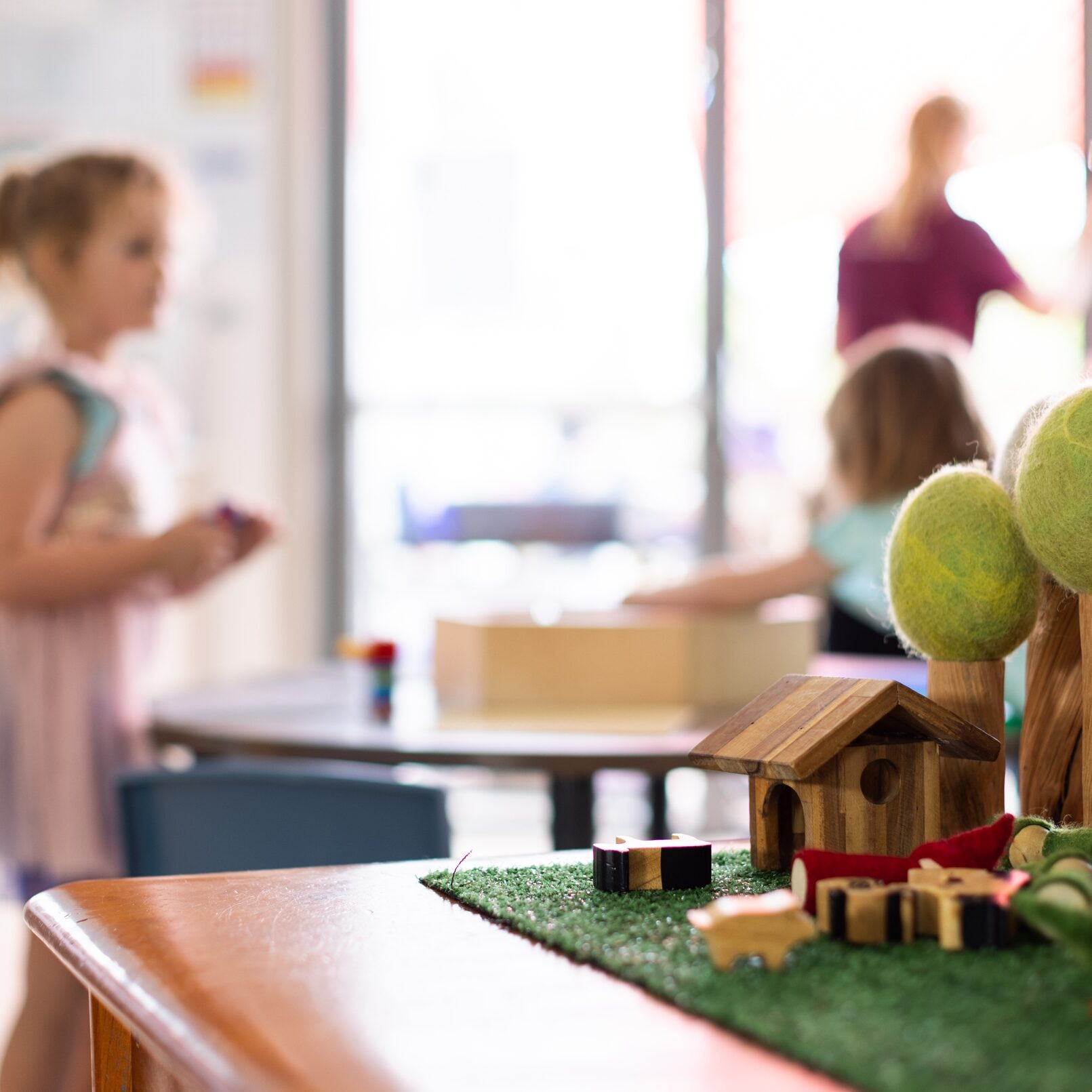Children playing in a bright classroom with toys on a table. Focus is on a wooden playset, while blurred background shows engaged kids and a caregiver. Warm atmosphere.