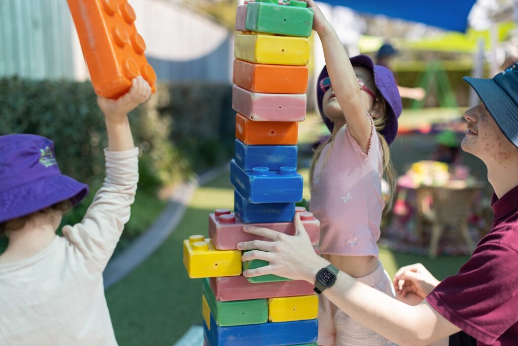 Tillys Children and an adult teacher play together, stacking large colorful blocks in a sunny outdoor area. The scene is lively and cooperative, emphasizing teamwork.