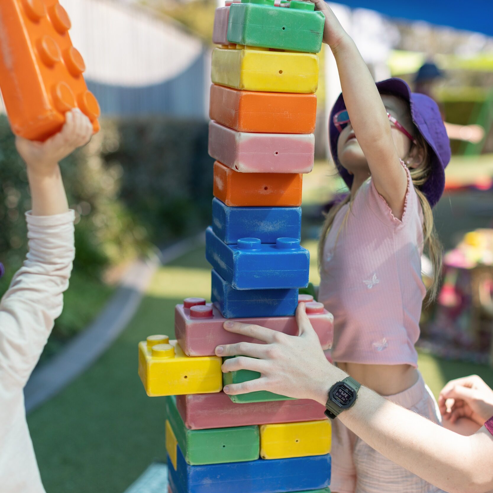 Children and an adult teacher play together, stacking large colorful blocks in a sunny outdoor area. The scene is lively and cooperative, emphasizing teamwork.