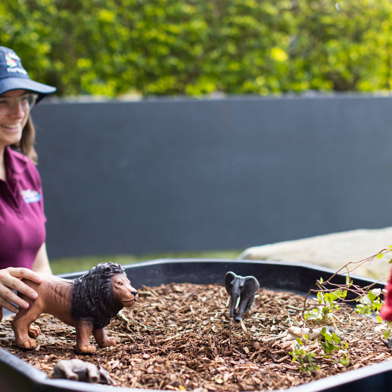A woman in a Tillys logo hat and glasses smiles, placing a plastic lion on a bed of mulch beside an elephant figurine. Greenery and a child are nearby as she watches the child play with a smile.