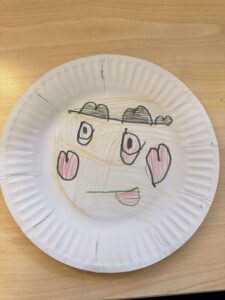 Child's drawing on a paper plate showing a face with heart-shaped eyes, a simple smile, cloud-like eyebrows, and a light brown background. Playful and whimsical.