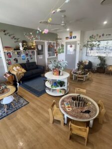 Bright, inviting playroom with wooden floors, small round tables, and chairs. Decorated with colorful hanging crafts and plants, creating a playful, cozy atmosphere.