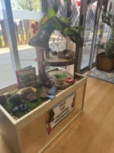 Wooden box shelf with a miniature treehouse labeled "Tree Top Treasure Treehouse." It features toy insects, a "Brilliant Bugs" book, and Spring garden decorations, creating a whimsical, educational scene.