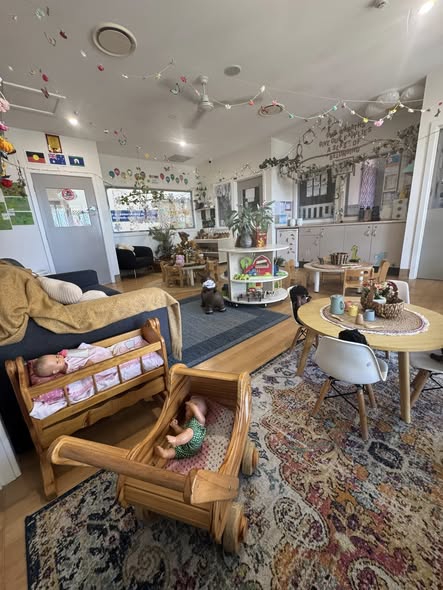 Cozy daycare room with toys, colorful garlands, and a rug. A wooden cradle and doll, round table with kids' chairs. Bright, welcoming atmosphere.