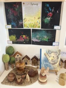 Children's art display featuring vibrant spring-themed paintings, a "How I Love You" book, wooden toys, and small house models on shelves, evoking creativity and warmth.