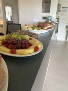 A table displays various platters of fruits, including grapes, pineapple slices, and raspberries, creating a vibrant and inviting afternoon tea spread in a modern room.