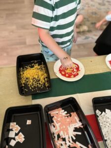 Tillys Rutherford A child in a green and white striped shirt arranges red and yellow paper bits on a plate to create a pizza collage. Black trays with colored paper shapes are on a tablecloth with green, white and red stripes.